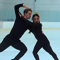 Evan Bates and Madison Chock in black costumes perform a dramatic dance pose on an indoor ice rink with glass panels and white walls in the background.