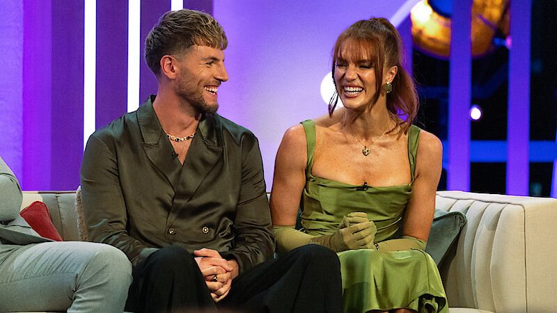 A man and woman sit closely on a couch, smiling and laughing in a brightly lit studio with vibrant purple lighting and a relaxed, cheerful atmosphere.