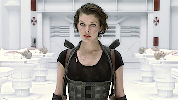 A woman in tactical gear stands in a futuristic, sterile laboratory with white walls and equipment, surrounded by examination tables holding covered bodies, suggesting a sci-fi or action setting.