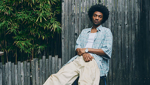 Jonathan Daviss with an afro hairstyle sits casually against a wooden fence outdoors, wearing a plaid shirt over a white t-shirt and cream pants, with green foliage in the background.