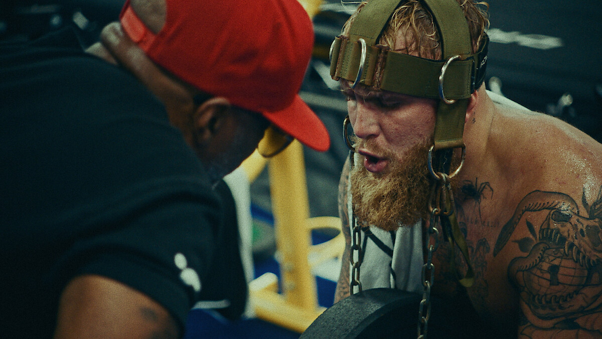 Jake Paul uses neck exercise equipment under the watchful eye of his trainer.