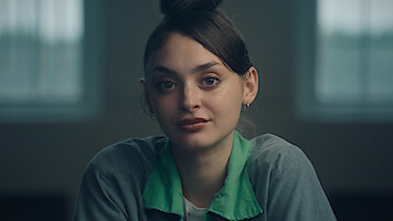 Young woman with dark hair in a bun, wearing a green-collared gray shirt, sitting indoors in a softly lit room with large windows in the background, looking directly at the camera with a calm expression.