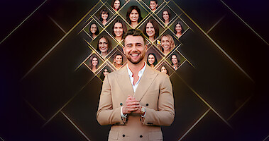 A smiling man in a tan suit stands in front of a dark background with glowing diamond shapes featuring multiple women’s faces displayed above and behind him, suggesting a reality TV or dating show setting.
