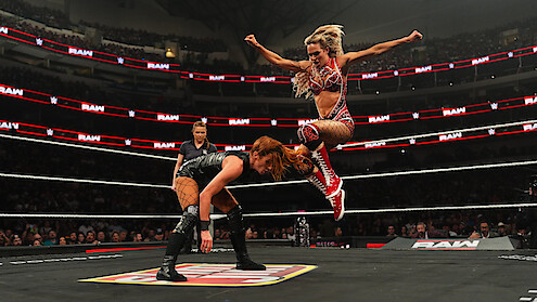 Female wrestler jumps toward opponent in a dramatic wrestling ring showdown with a referee and large cheering crowd in a stadium, vibrant lighting, and intense, energetic atmosphere.