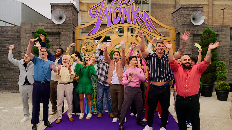 A diverse group of people cheering joyfully outside a large decorative golden gate with the word "Wonka" above it, set in an outdoor area with potted plants and brick buildings in the background.