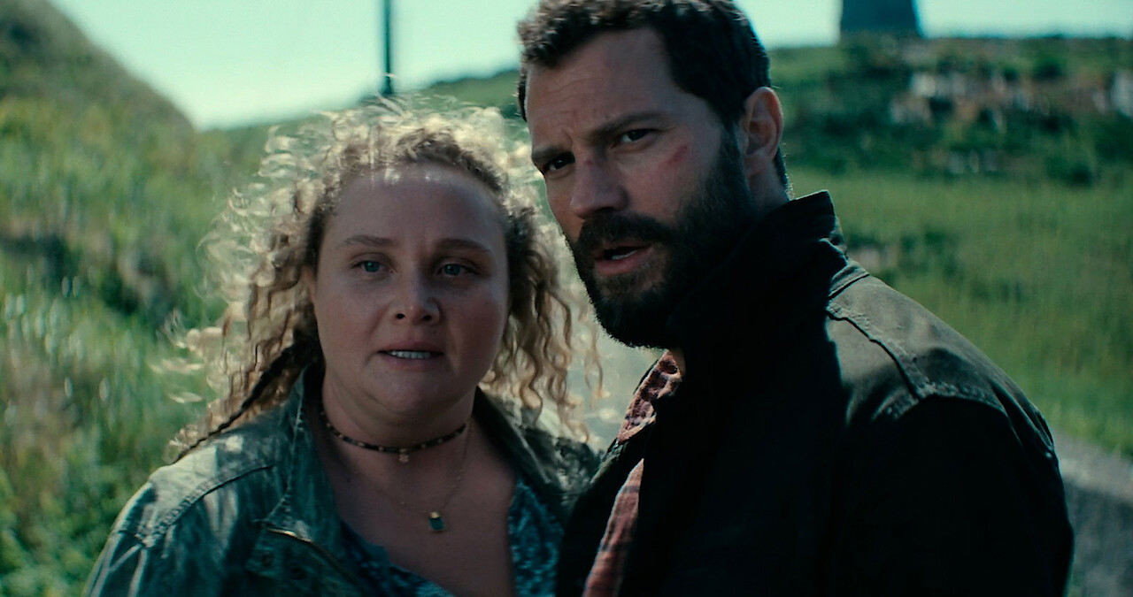 Jamie Dornan as Elliot and Danielle Macdonald as Helen stand together on a grassy hill in season 2 of 'The Tourist'