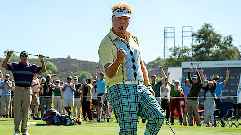 Golfer celebrating on green with fist pump, wearing plaid pants and visor, as cheering crowd watches in a sunny outdoor golf course setting with scoreboard and hills in background.