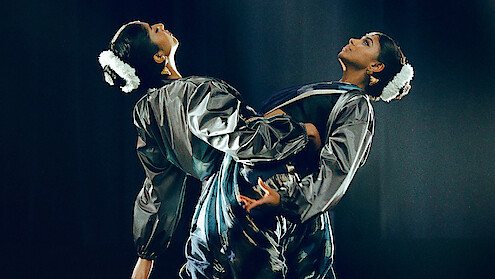 Two dancers in matching shiny costumes perform on stage with dramatic lighting, facing away from each other and leaning back-to-back, wearing white floral hair accessories in a dark, theatrical setting.