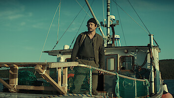 A man wearing a beanie stands on the deck of a fishing boat surrounded by nets and wooden railings, with the sea and distant land visible under a clear sky.