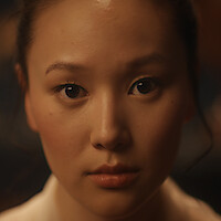 Close-up portrait of a woman with a neutral expression, softly lit in a warm indoor environment, dark blurred background.