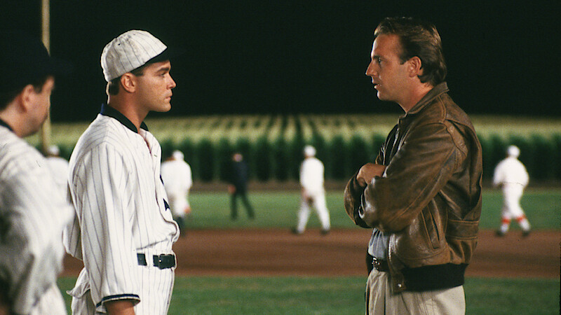 Two men stand on a baseball field at night, one in a vintage striped uniform and the other in a brown jacket, talking seriously; players and a cornfield are visible in the background under stadium lights.