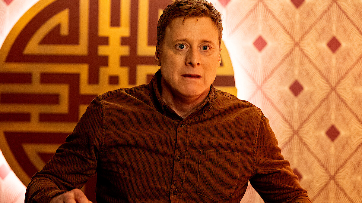 A man in a brown shirt looks surprised in a warmly lit room with patterned wallpaper and a large decorative circular wall design behind him.