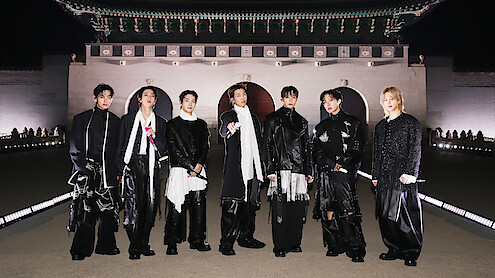 V, JIN, SUGA, RM, JUNG KOOK, J-HOPE, and JIMIN dressed in fashionable black and white outfits stand outdoors at night in front of an ornate, traditional East Asian building with arches and a large decorative roof.