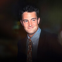 A young man in a suit and patterned tie smiles slightly, standing in a dimly lit environment with soft, colorful bokeh lights in the background, suggesting an evening or nighttime urban setting.