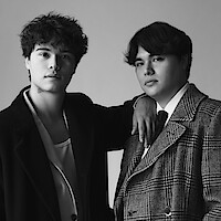Two young men pose together in a studio, one with his arm resting on the other's shoulder. Both wear stylish coats, one in a suit and tie, in a black and white portrait with a plain background.