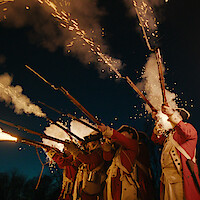 Historical reenactors in red coats fire muskets at night, with sparks and smoke lighting up the dark sky, evoking a dramatic battle scene from the 18th century.