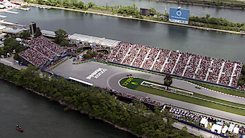 Aerial view of a Formula 1 racetrack section surrounded by water, grandstands filled with spectators, green trees, and a large billboard visible across the water.