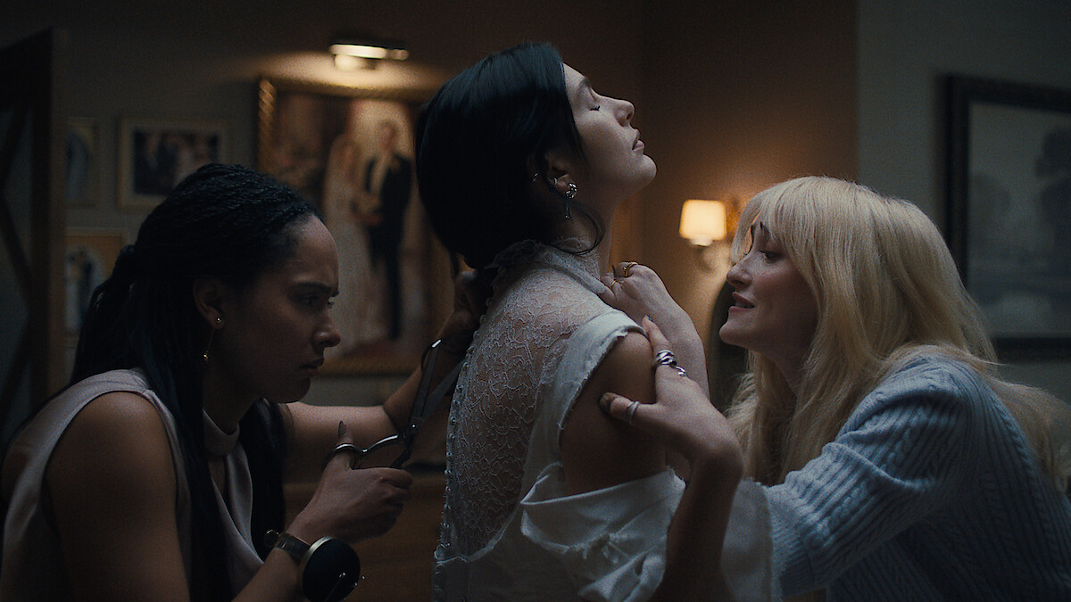 Three women in a dimly lit room, one holding scissors cut the back of another’s white lace dress while a third woman appears to comfort or steady her, with framed art and soft lamp lighting in the background.