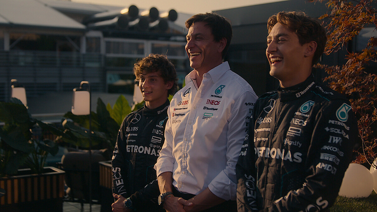 Three people, two in black Mercedes Formula 1 racing suits and one in a white team shirt, stand smiling outdoors at a race venue with plants and a modern building in the background during daylight.