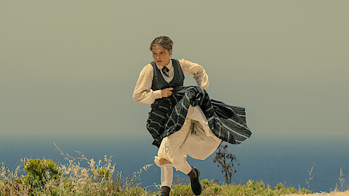 Woman in period clothing running outdoors on a grassy hill with the ocean and clear sky in the background.