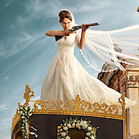 Bride in a white wedding gown stands on a decorated carriage, aiming a rifle, with a flowing veil and historic building in the background under a blue sky.