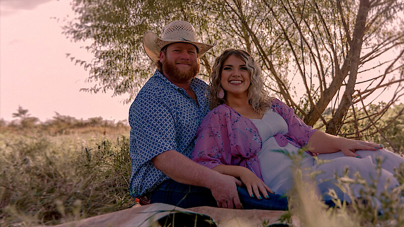 Smiling couple sitting on a blanket outdoors in a grassy field, with trees in the background, wearing casual clothes and cowboy hat, enjoying a relaxed moment in nature during daylight.