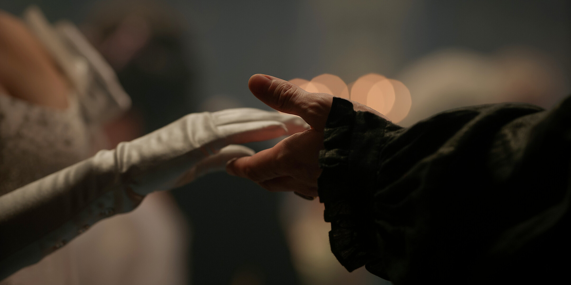 Two people reach toward each other, one wearing a white glove and elegant attire, the other in dark clothing, set in a softly lit, formal ballroom with blurred figures and warm lights in the background.