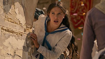Young woman with long hair in historic clothing leans anxiously around a stone wall in an outdoor setting, with colorful confetti in the air and other people nearby, suggesting a dramatic or festive event.