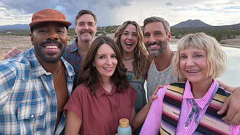Group of six smiling adults taking a selfie outdoors by a pool, with mountains and cloudy sky in the background, casual clothing, relaxed atmosphere, desert landscape.