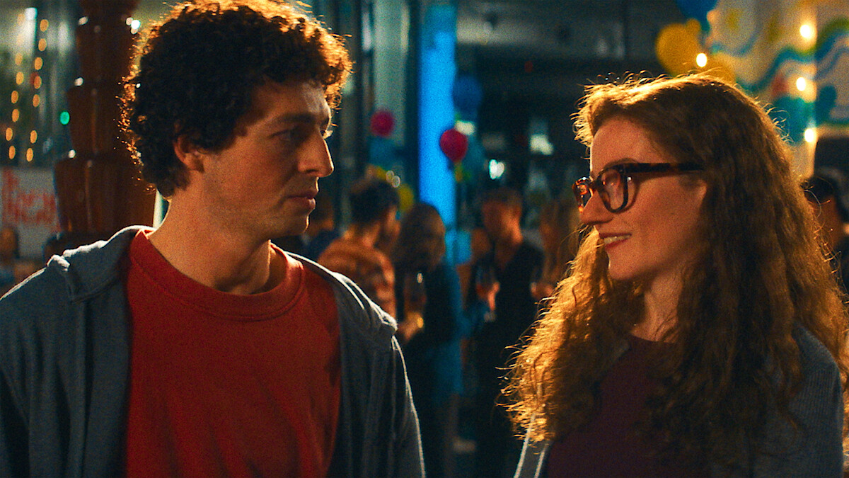Two young adults, a man with curly hair and a woman with glasses, smile and talk at a festive indoor party decorated with balloons and lights, surrounded by other people socializing in the background.