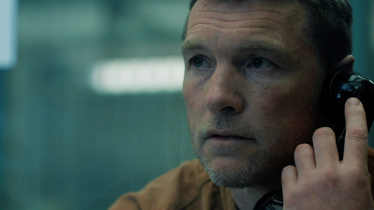 Man with short hair and stubble sits indoors, holding a black phone receiver to his ear, looking serious. The background is blurred, suggesting he is in a visitation booth or interrogation room, likely in a prison or secure facility.