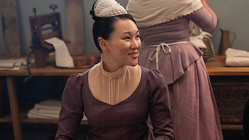 A woman in a purple historical dress and cap sits smiling in a rustic room with wooden tables, linens, and vintage laundry tools, while another similarly dressed woman works in the background.