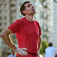 Alex Honnold in a red shirt stands outdoors in an urban setting, looking upwards with a thoughtful expression. Blurred tall buildings and trees are in the background, suggesting a city environment.