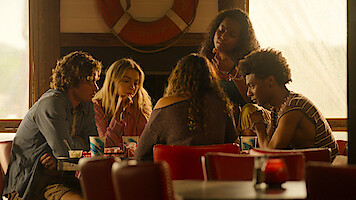 Five young people sit around a table in a retro diner with red chairs and nautical decor, talking and sharing drinks, sunlight streaming through the windows, creating a warm, casual and friendly atmosphere.