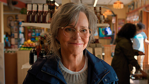 Smiling older woman with gray hair and glasses in a blue jacket stands inside a well-lit grocery store, with shelves of food and drinks, people shopping, and produce in the background.