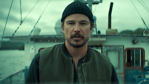 A man wearing a black beanie and green vest stands on a fishing boat, with water and cloudy sky in the background, conveying a rugged, maritime setting.