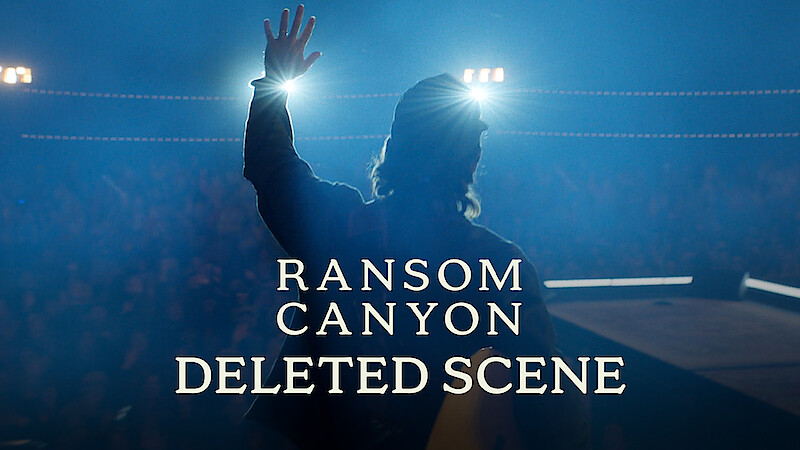 Thomas Rhett stands on stage in silhouette, facing bright spotlights with one hand raised. Smoky blue lighting fills the scene, the audience is barely visible in the background, and a text overlay reads: "Ransom Canyon Deleted Scene."