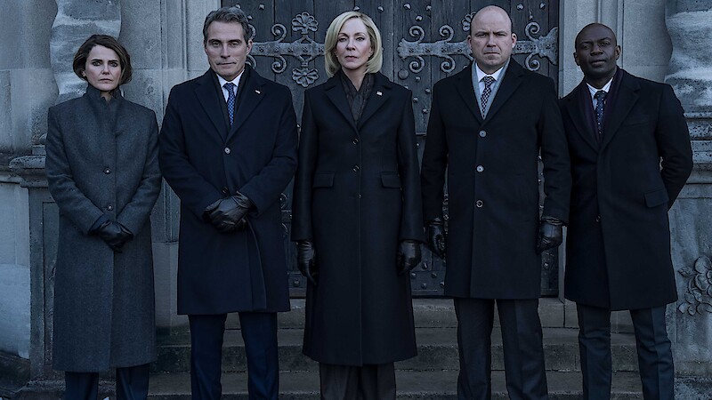 Three people in dark formal coats and gloves stand outside in front of an old stone building and ornate doors, posing seriously for a photograph in a somber or official setting.
