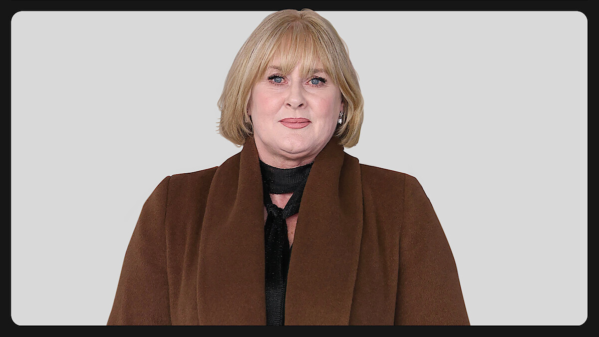 A person with short blonde hair wearing a brown coat and black top stands against a plain light gray background, facing the camera with a neutral expression.