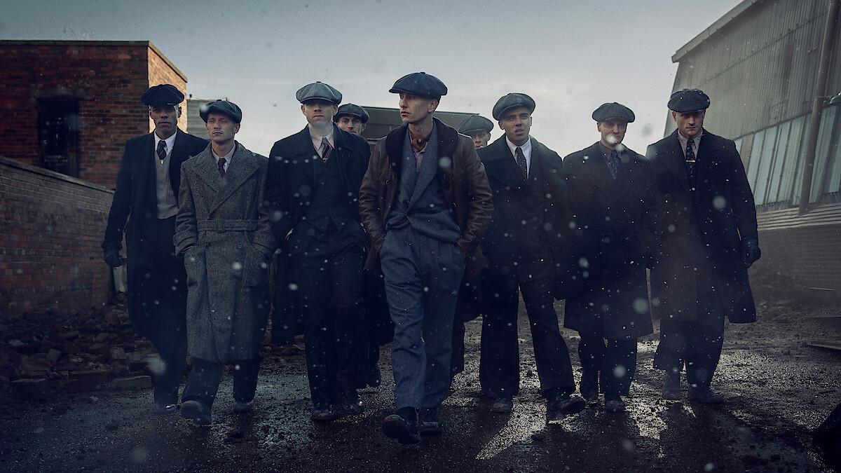 A group of men in vintage suits and newsboy caps walk together closely on a rainy, industrial street. The mood is serious and gritty, with cold light and falling rain enhancing the dramatic, early 20th-century atmosphere.