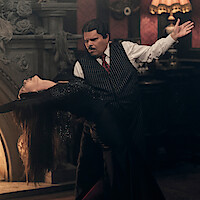 Catherine Zeta-Jones as Morticia Addams and Luis Guzmán as Gomez Addams dancing together in front of a fireplace in 'Wednesday' Season 2.