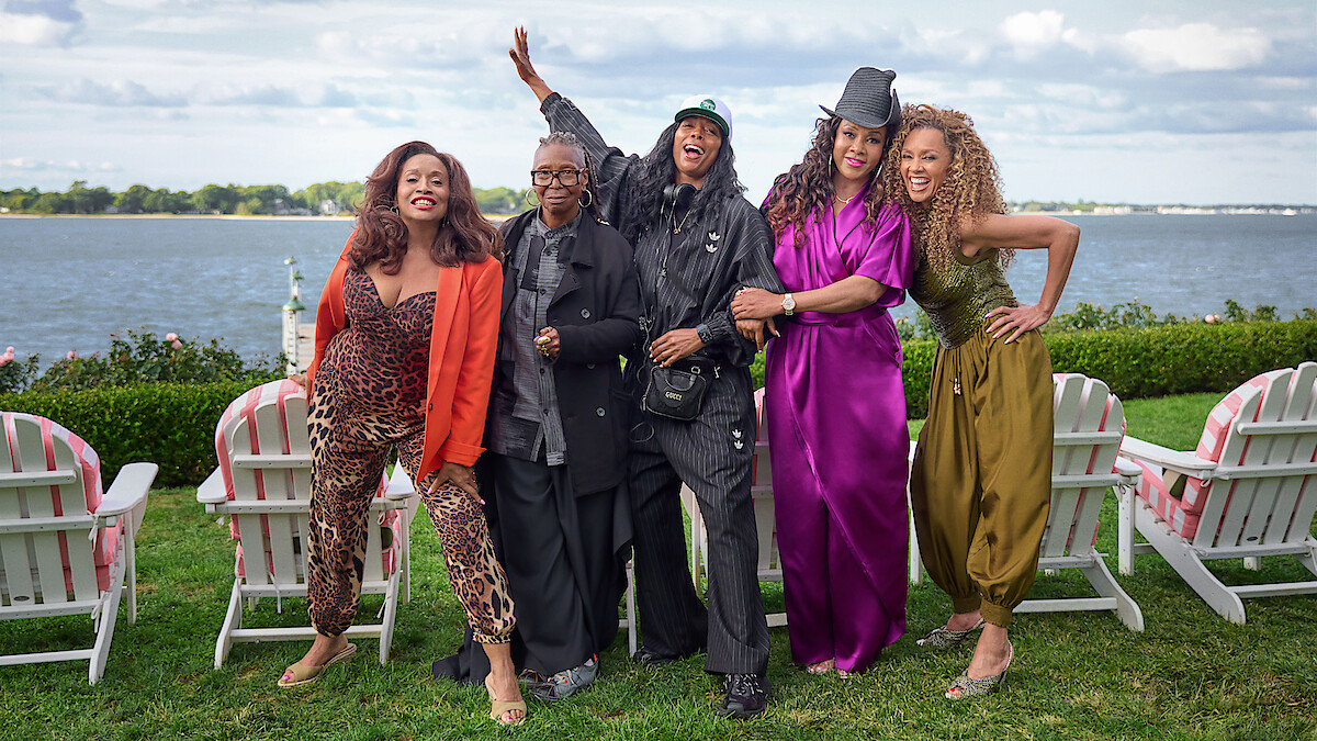 Five stylish women stand outdoors on green grass in front of white chairs by a lake, smiling and posing happily under a partly cloudy sky.