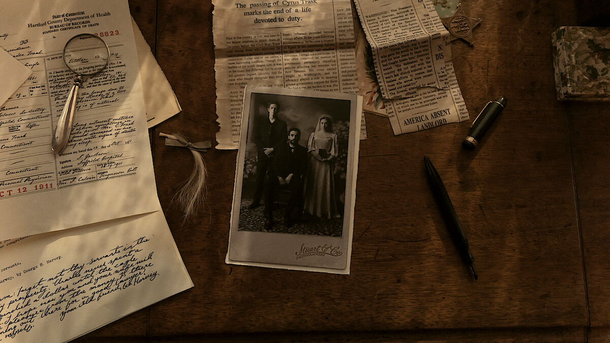 Old sepia photograph, handwritten letters, magnifying glass, pen, and newspaper clippings spread out on a wooden desk, suggesting investigation or family history research in a vintage setting.