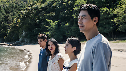 Four people stand side by side on a sandy beach with trees and greenery in the background, looking thoughtful under natural daylight by the shoreline.