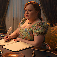 Woman in a floral dress writes with a quill at an ornate wooden desk with candles, in a lavish, candlelit room with elegant curtains and antique decor.