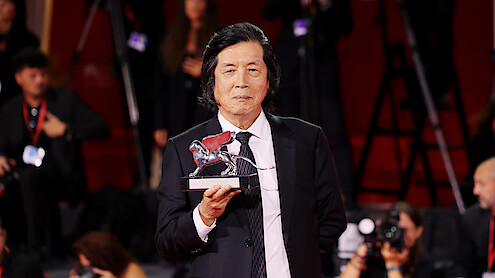 Man in a suit holding a silver lion trophy on a red carpet, surrounded by photographers and people with cameras and badges in an event or awards ceremony setting.