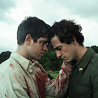 Two injured men in bloodstained uniforms comfort each other, standing close with foreheads touching outdoors under a cloudy sky, conveying emotion and solidarity in a natural, green environment.
