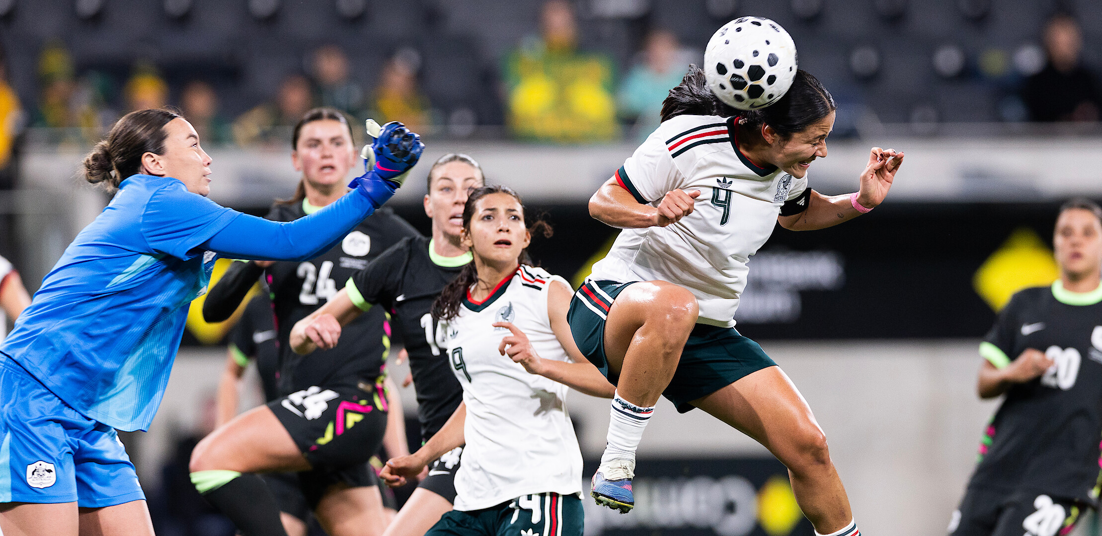 Women's soccer match in a stadium, goalkeeper in blue reaches for the ball as player in white and green jumps to head it, teammates and opponents in action, focused expressions, blurred spectators in background.