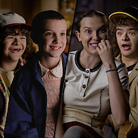Gaten Matarazzo and Millie Bobby Brown from Stranger Things, shown as both kids and adults, seated in interview settings, reacting and smiling.