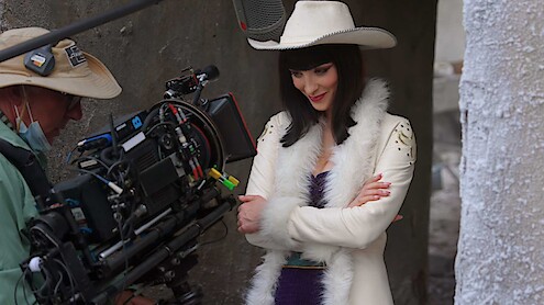 A woman in a white fur coat and cowboy hat poses on a film set, smiling with arms crossed, while a cameraman in outdoor gear films her in a narrow concrete corridor.